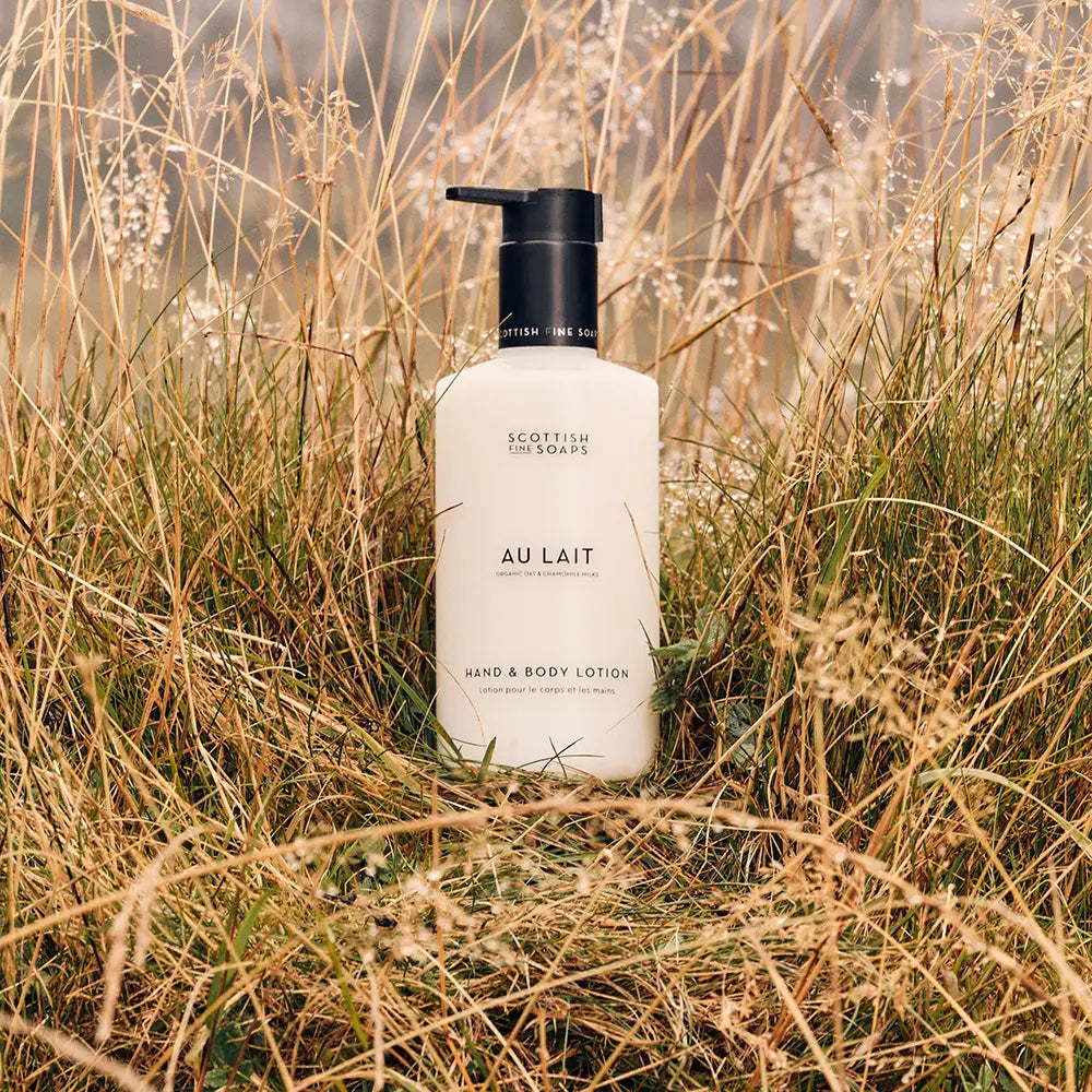 A bottle of Scottish Fine Soaps 'Au Lait' hand and body lotion, featuring a matte white bottle and a black pump dispenser, in a grassy field