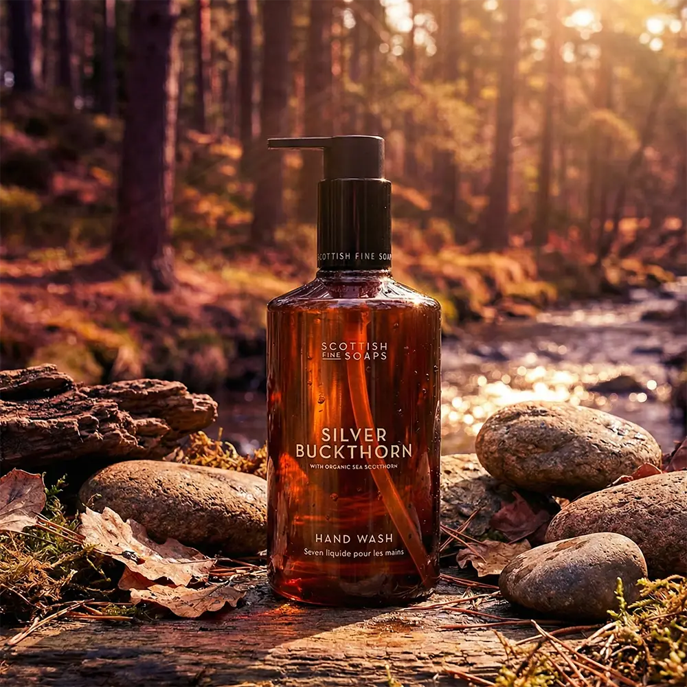 A bottle of Scottish Fine Soaps Silver Buckthorn Hand Wash stands on a log in a sunlit forest stream setting. The amber bottle has a black pump dispenser and features white text, set against a blurred background of trees and water.