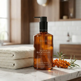 A bottle of Scottish Fine Soaps Silver Buckthorn hand wash stands on a marble countertop in a softly blurred bathroom setting, with folded towels on the left and a small cluster of orange sea buckthorn berries on the right.