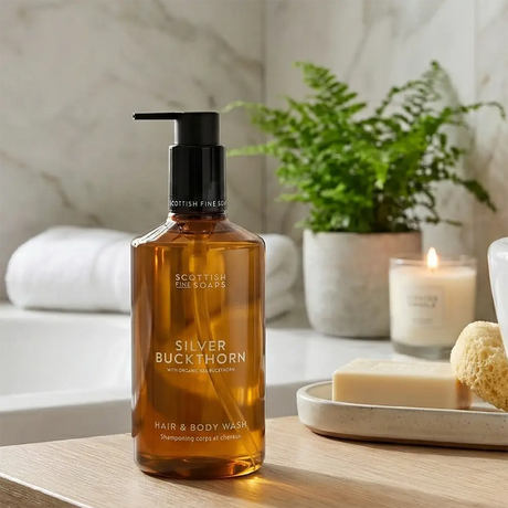 A bottle of Scottish Fine Soaps Silver Buckthorn hair and body wash stands on a bathroom counter, with a white tub, folded towels, a potted fern, a lit candle, and a sponge in the background.