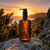 A bottle of Scottish Fine Soaps Silver Buckthorn Hair & Body Wash stands on a rock outdoors at sunset, with mountains and trees in the background.