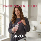 Woman pouring a Spruce Raspberry & Elderflower sachet into a Spruce water bottle in a kitchen, 'Bring Water To Life' tagline