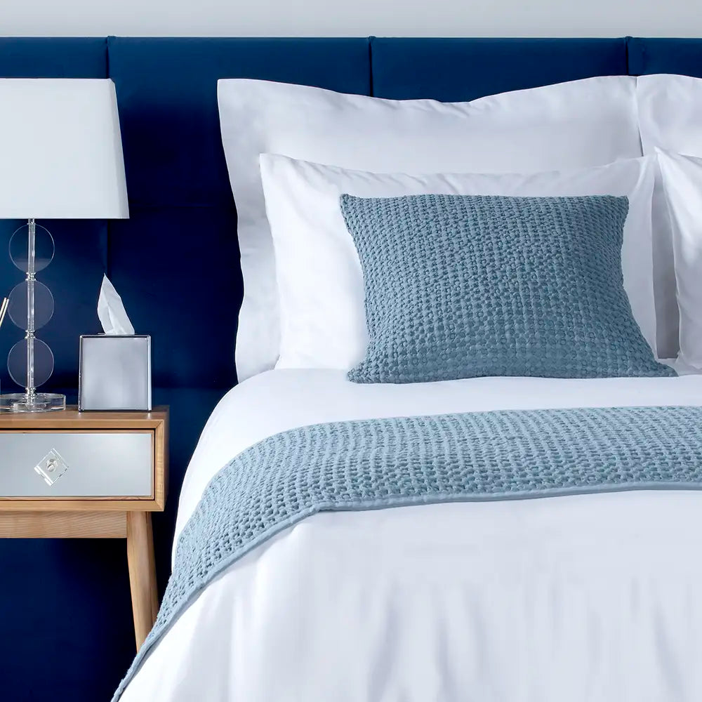 Corded Oxford bedsheets set on a blue bed frame featuring a blue textured throw, nightstand with lamp and tissue box