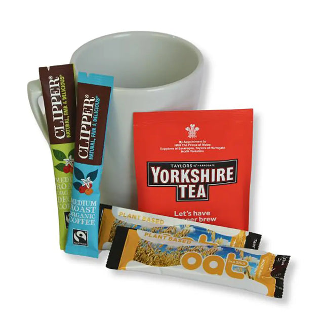 White mug with Clipper coffee packets, Yorkshire Tea packet, and Oatly oat milk stick on a white background