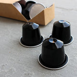 Wheatleys Fairtrade Colombian Arabica Coffee Pods - Pack of 120 -Three black coffee capsules on a concrete surface with a cardboard box in the background.