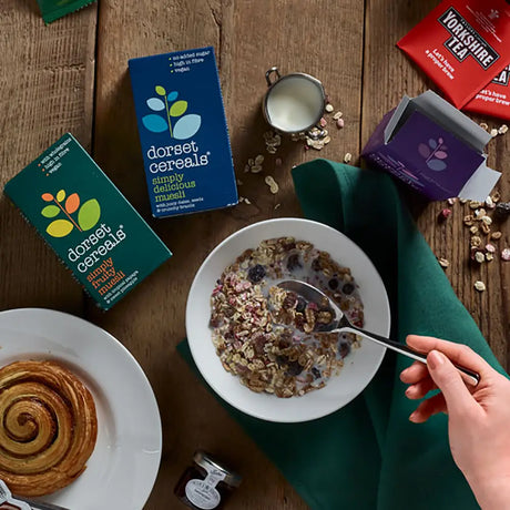Breakfast scene with Dorset Cereals Simply Delicious Muesli Portion Packs cereal bowl, croissant, and tea on a wooden table.
