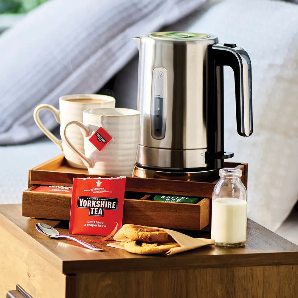 Electric kettle on a wooden tray with yorkshire tea bags, milk, and biscuits on a wooden surface.