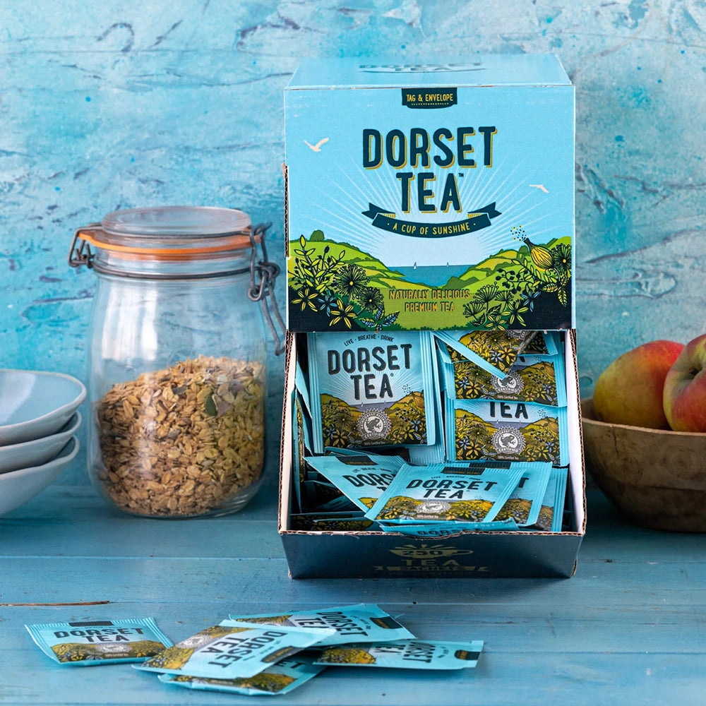 Box of Dorset Tea on a blue wooden surface with a jar and apples in the background.