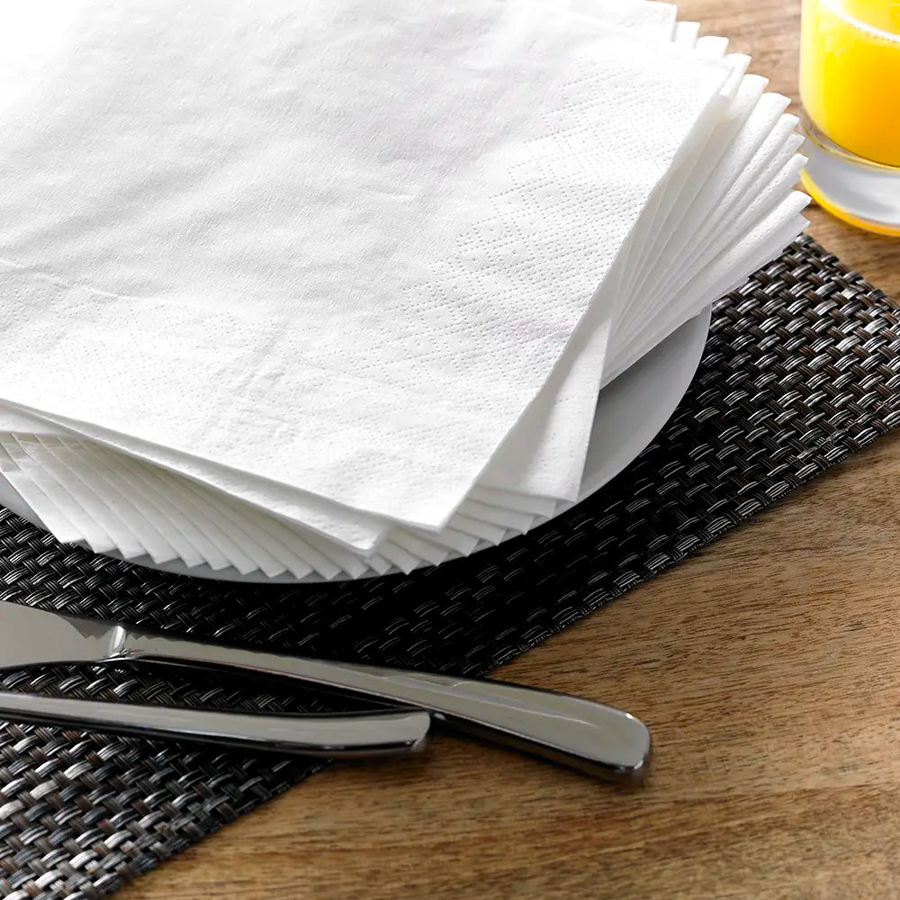 Neat pile of 2 ply paper napkins on a woven placemat with silverware and a yellow glass in the background.